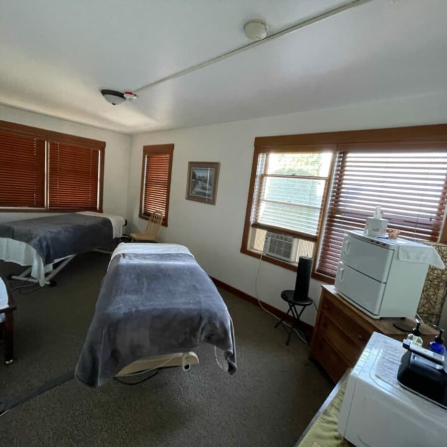 A calm massage room featuring two treatment tables, a chair, windows with wooden blinds, and a small side table with equipment.