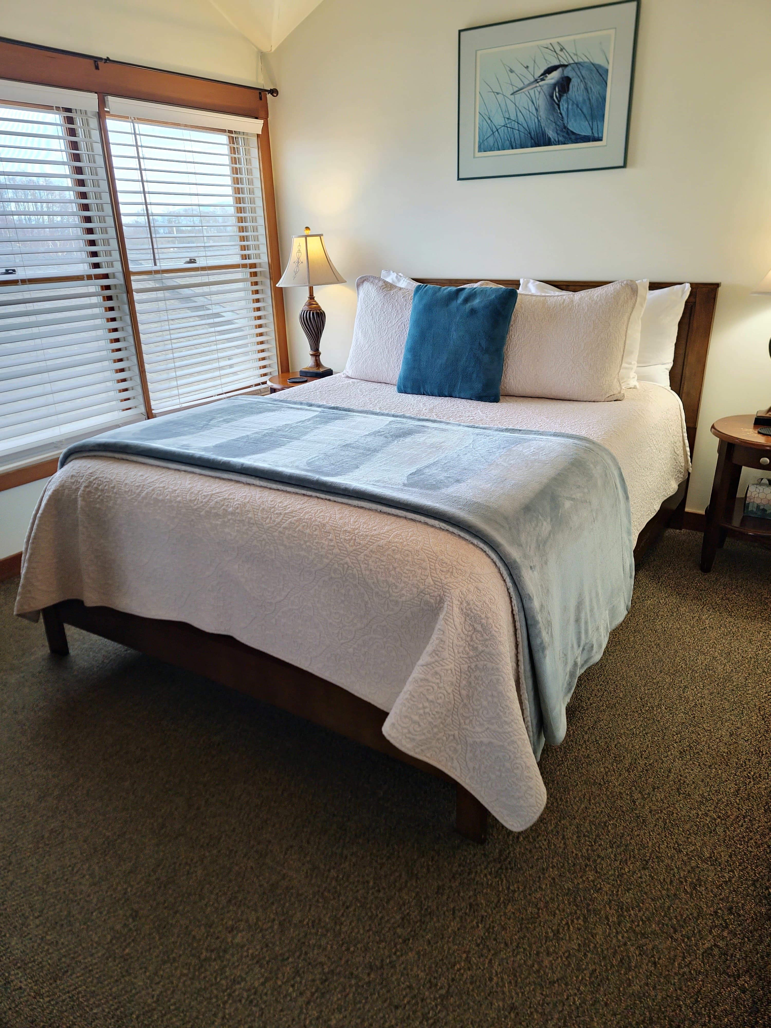 A cozy bedroom featuring a double bed with plush pillows, a light blue blanket, and a bedside lamp beside a window.