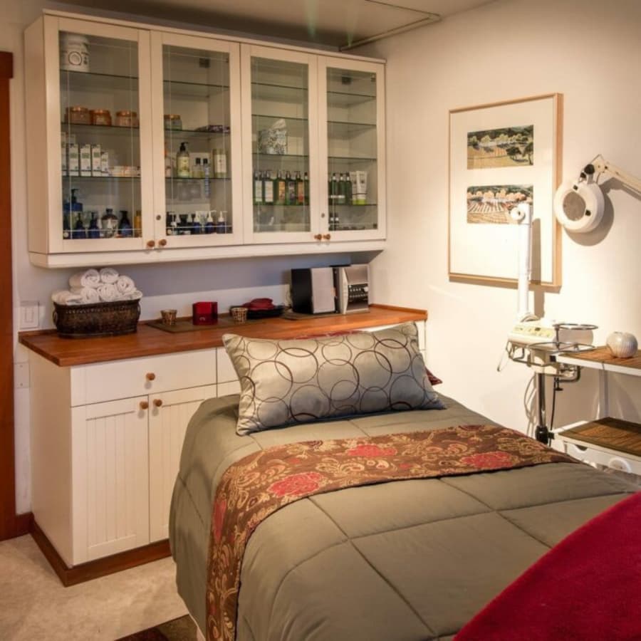 A cozy treatment room featuring a bed with decorative bedding, a glass cabinet with various skincare products, and massage tools.