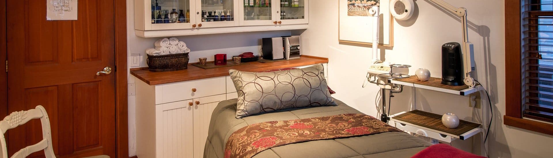 A cozy treatment room featuring a massage bed, storage shelves, and warm lighting.