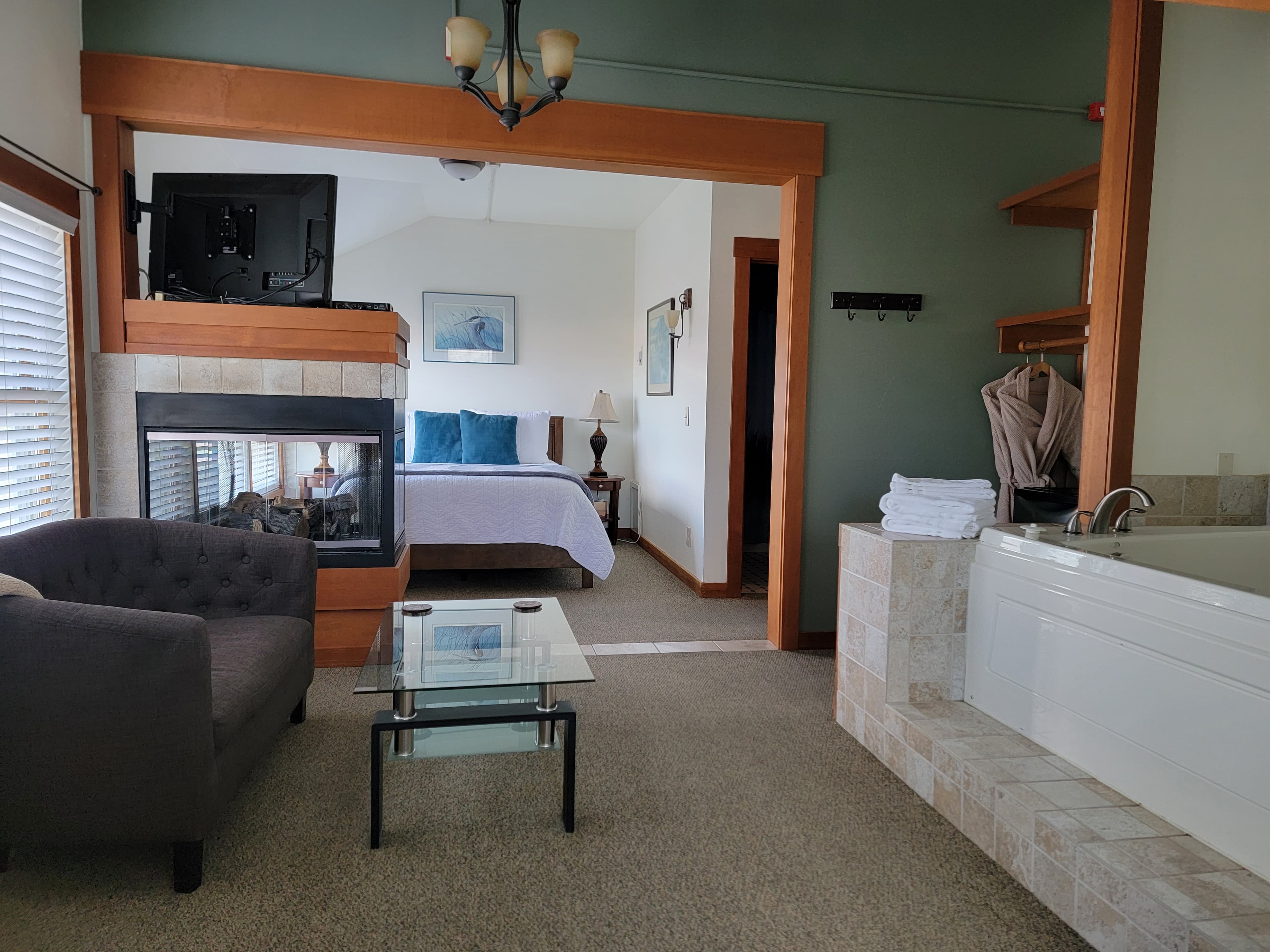 A cozy hotel room featuring a sitting area with a gray couch, a glass coffee table, a fireplace, and a king-sized bed in the background.