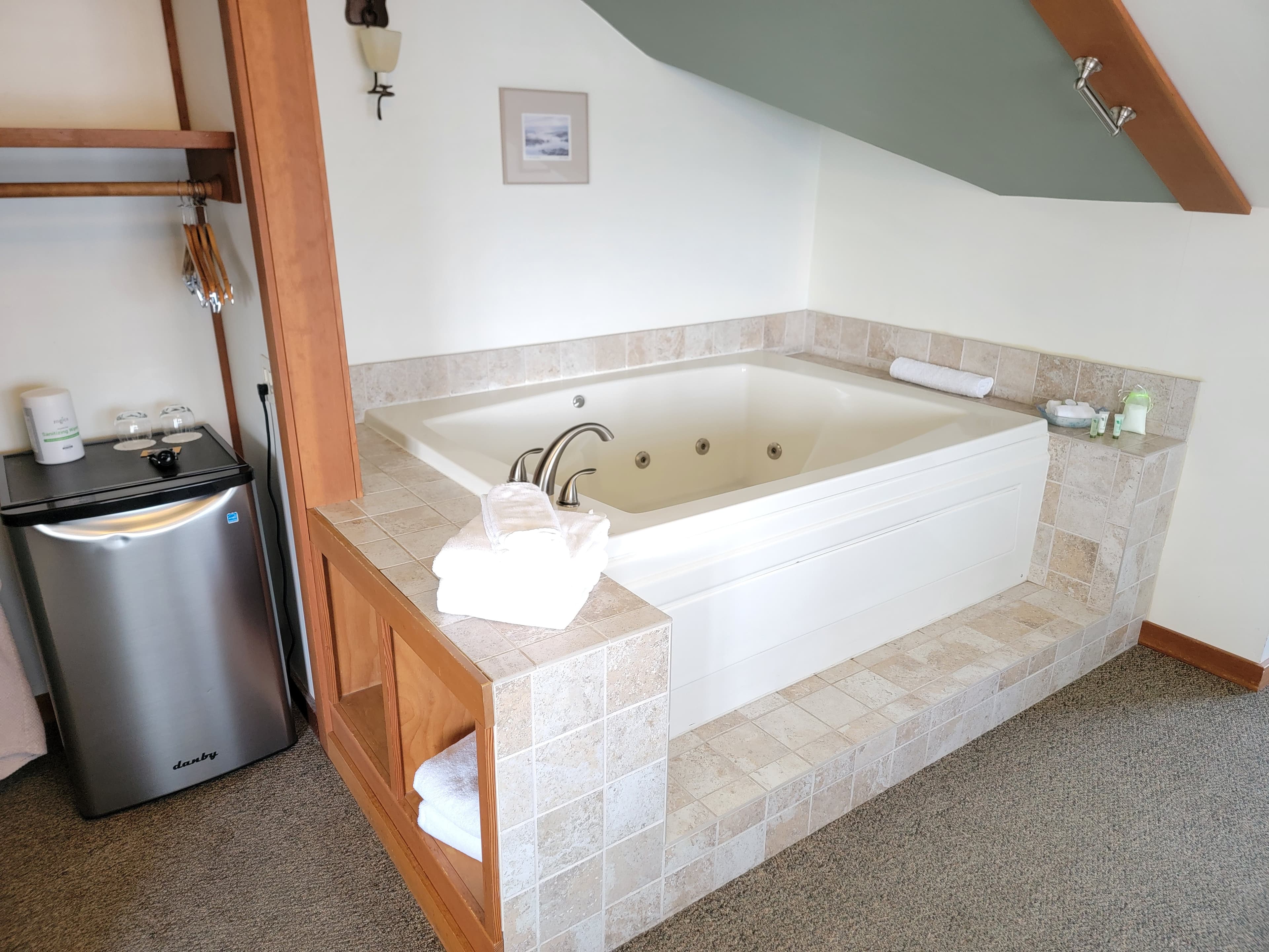 A spacious bathroom featuring a white jacuzzi tub, a small fridge, and a neatly arranged towel display.